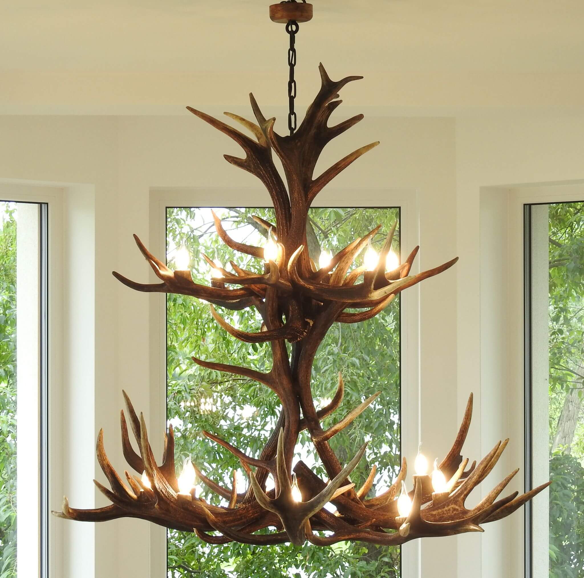 Farmhouse antler chandelier hanging in a room with large windows showing greenery outside.